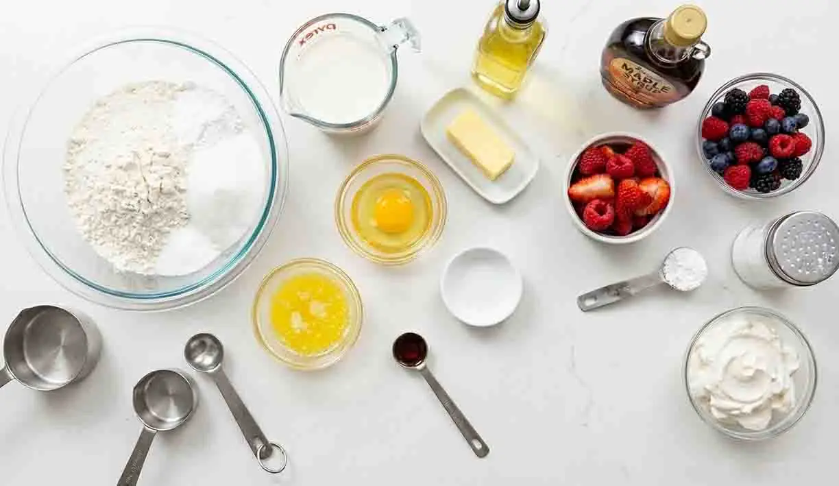 Fluffy Pancakes Delight: A Heavenly Breakfast Treat! 2 Fluffy pancakes ingredients arranged on a table including flour, eggs, milk, butter, maple syrup, and fresh berries