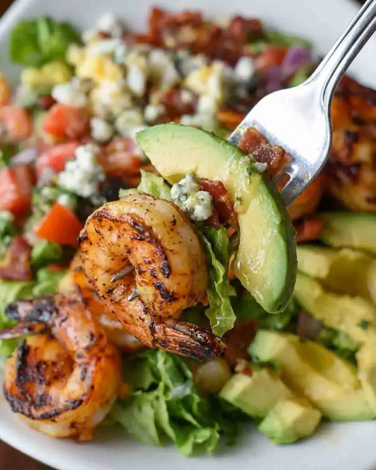Grilled Shrimp Avocado Salad: A Burst of Zesty Freshness 3 Fork holding grilled shrimp and avocado slice over a fresh shrimp avocado salad with lettuce, tomatoes, and crumbled cheese