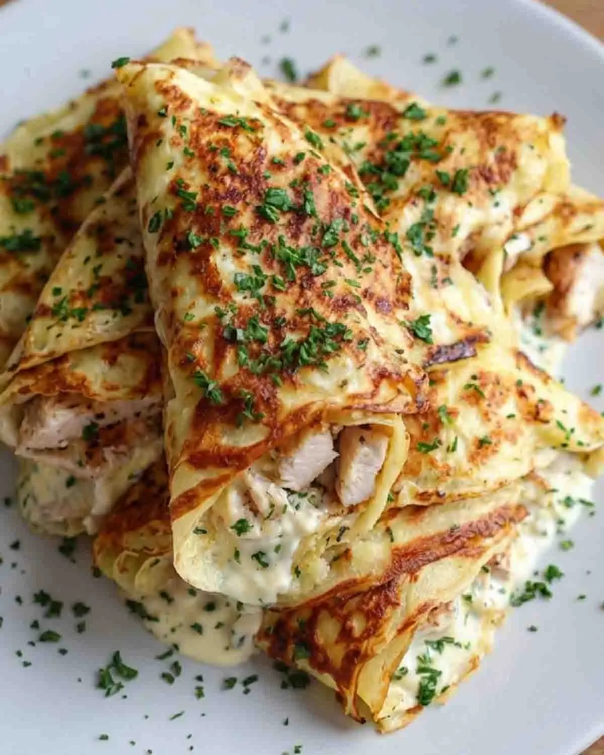 Savory Crepe Indulgence: Chicken Crepe Wraps for a Flavorful Lunch! 3 Golden chicken crepe wraps filled with creamy chicken and herbs, folded and stacked with a rich sauce and sprinkled with fresh parsley