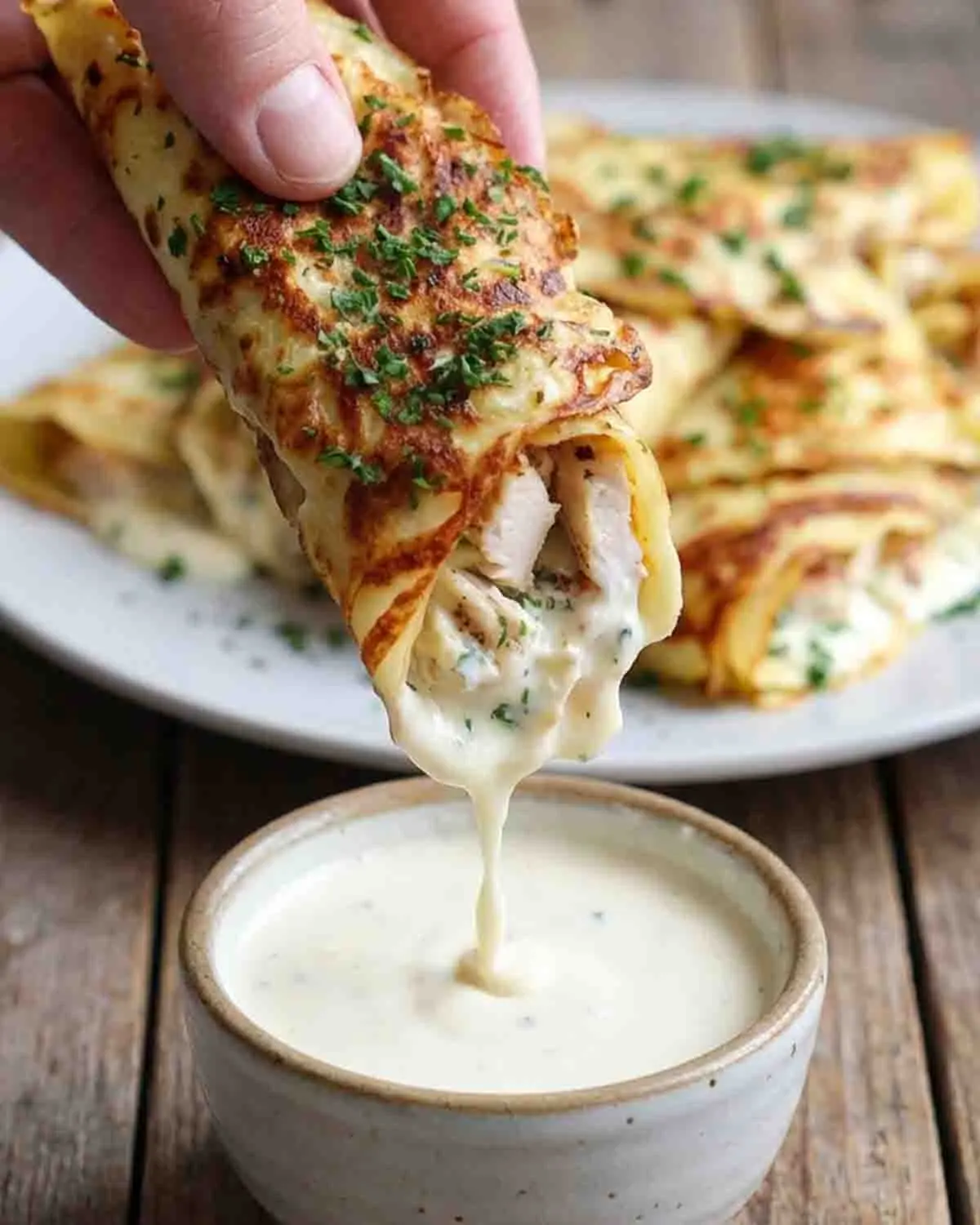 Savory Crepe Indulgence: Chicken Crepe Wraps for a Flavorful Lunch! 4 Hand holding a golden chicken crepe wrap filled with creamy chicken, dipping into a rich garlic cream sauce with herbs