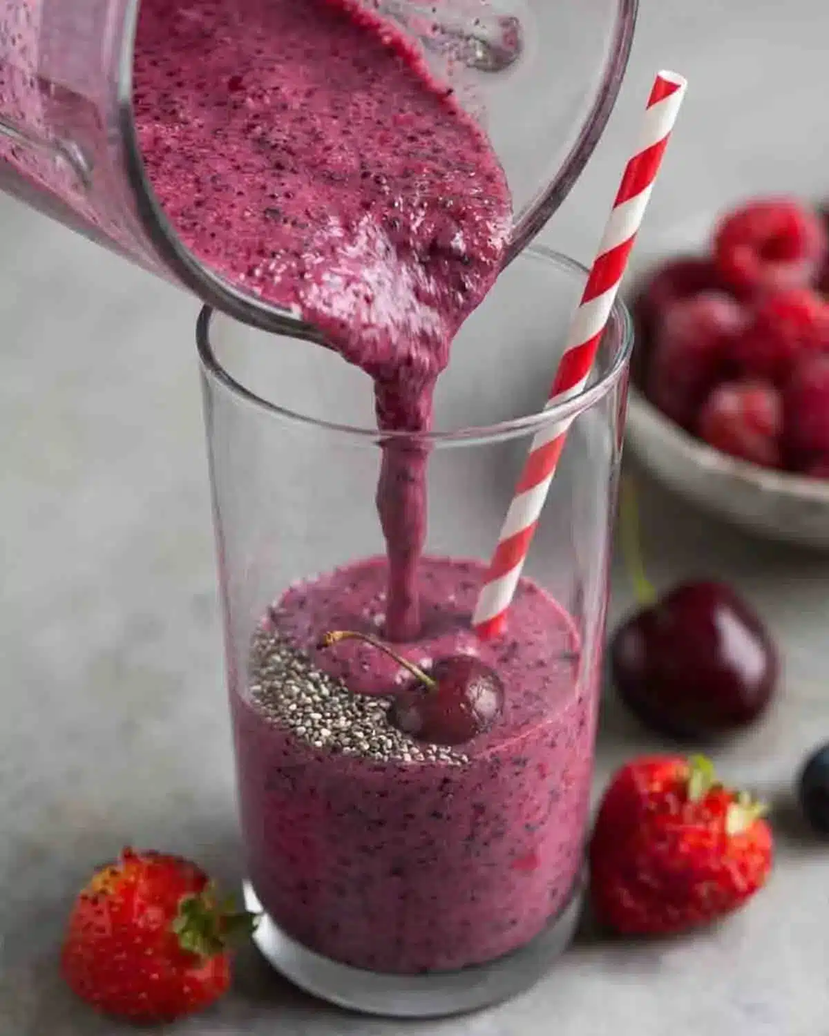 Berry, Chia, and Mint Smoothie 3 Berry chia and mint smoothie being poured into a glass with chia seeds and fresh cherries, surrounded by strawberries and mixed berries