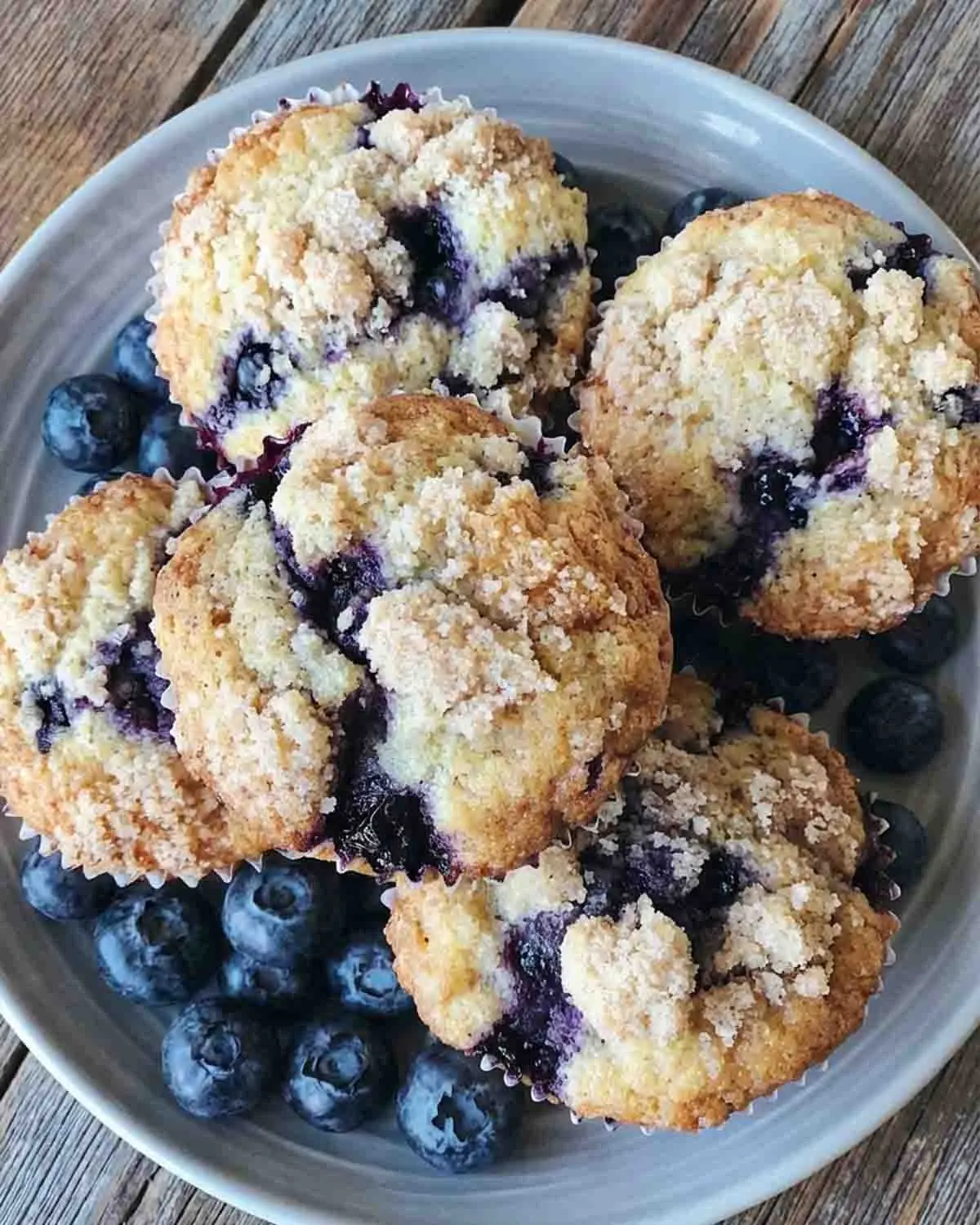 Blueberry Muffins Recipe 4 Plate of homemade blueberry muffins Recipe with crumb topping, golden brown and bursting with fresh blueberries, served with whole blueberries on a rustic wooden table.