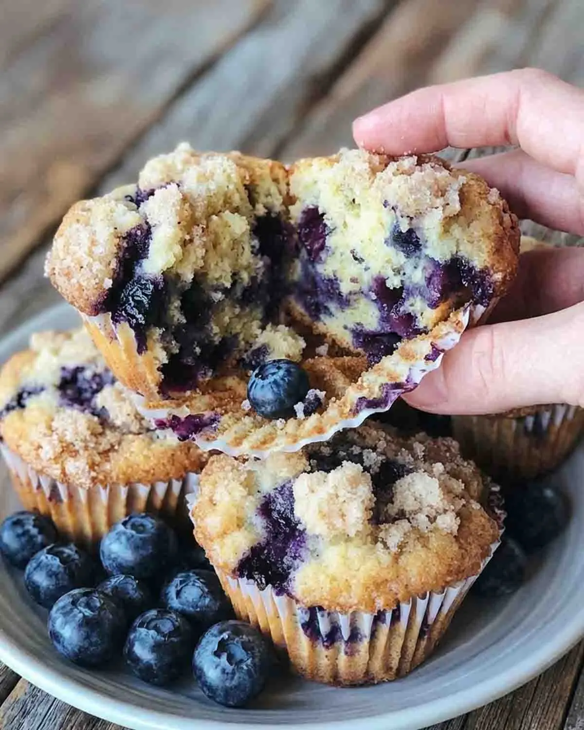Blueberry Muffins Recipe 3 Freshly baked blueberry muffins with crumb topping and juicy blueberries, partially visible on a plate in a kitchen setting.