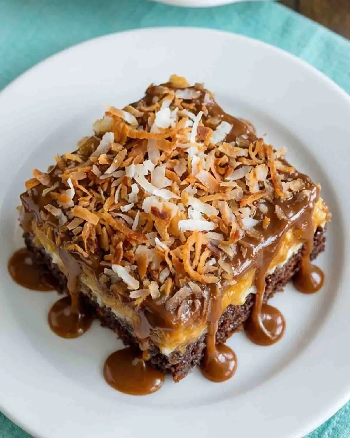 German Chocolate Poke Cake 3 German Chocolate Poke Cake slice topped with toasted coconut, pecans, and caramel drizzle on a white plate, showing moist chocolate layers and creamy filling.