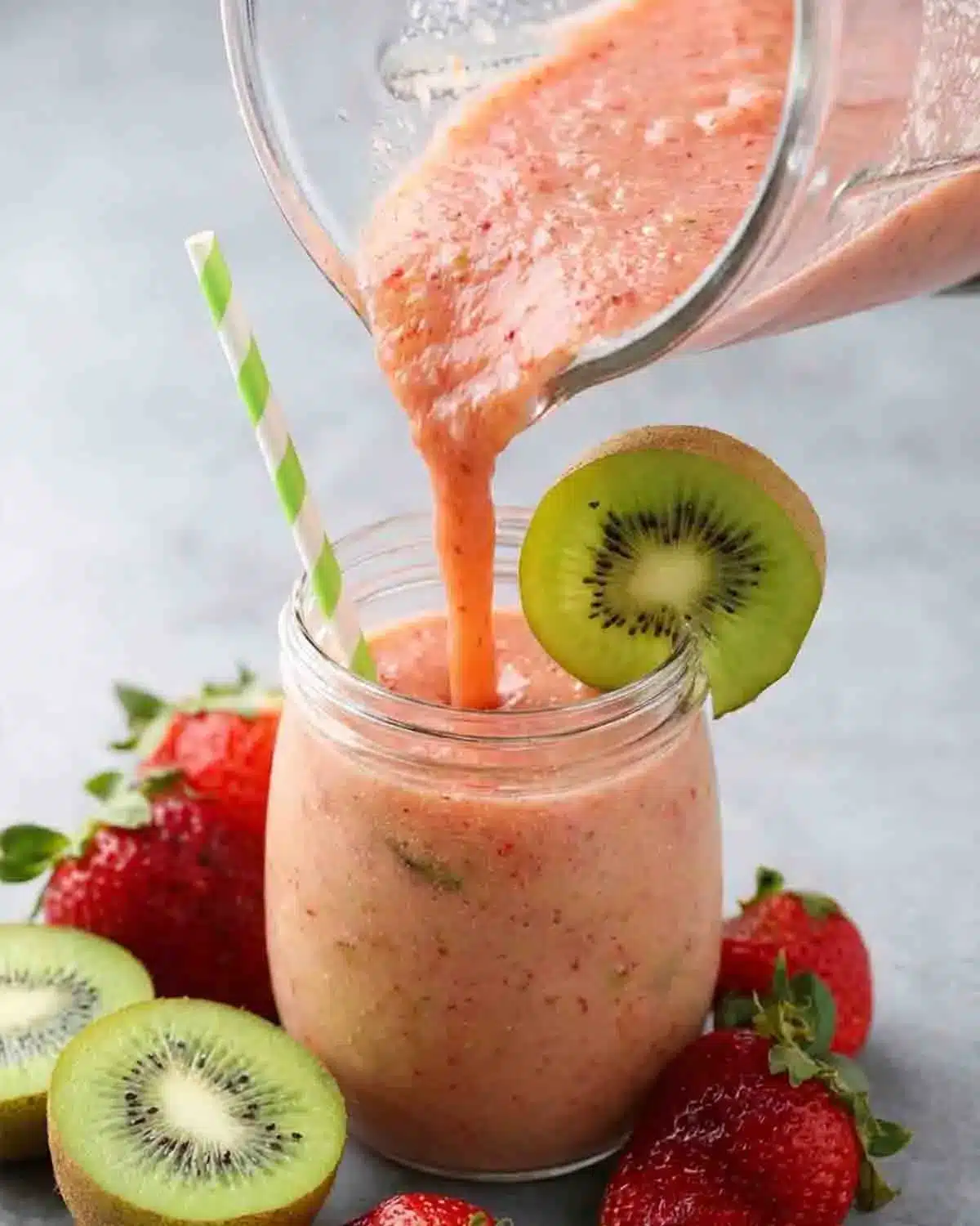 Strawberry-Kiwi Smoothie 4 Pouring Strawberry-Kiwi Smoothie into a glass jar with fresh kiwi slice garnish and strawberries on the side